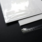 A close-up of a clear, glossy sheet of material placed over a white sheet on a dark black background, with a separate strip of the same transparent material shown in front.