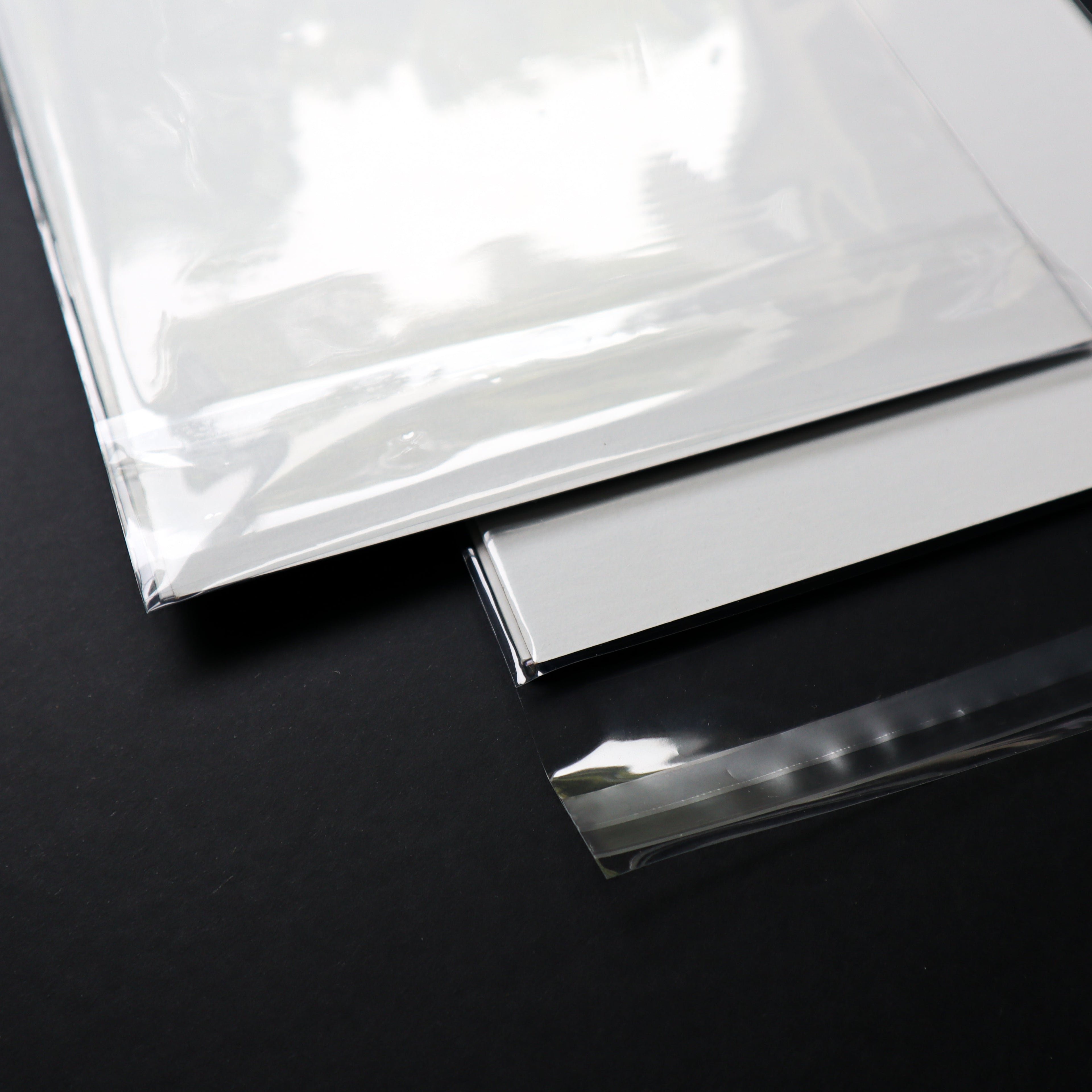 A close-up of a clear, glossy sheet of material placed over a white sheet on a dark black background, with a separate strip of the same transparent material shown in front.